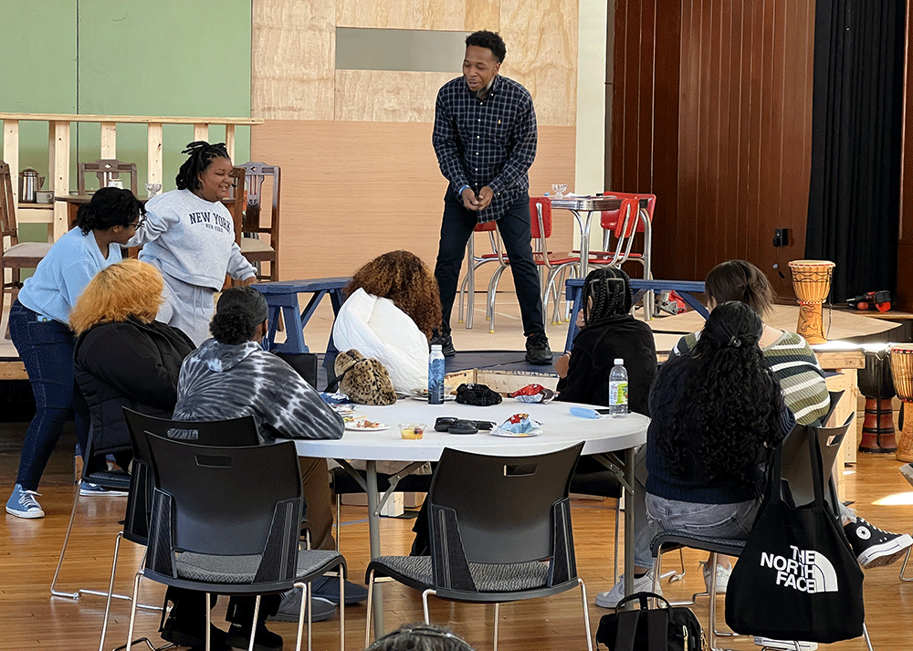 Seven high school students seated at round table receiving directions from instructor